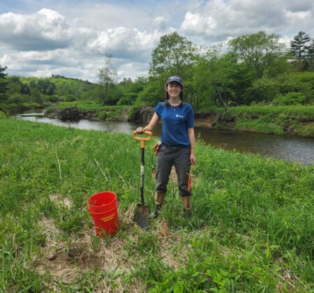 Woman riparian planting trees at West River with bucket and shovel