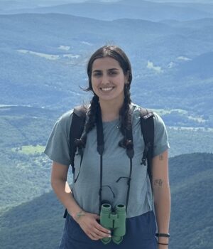 Gabriella Marchesani, a UVM and University of Florida alumna, stands on Camel’s Hump Mountain holding binoculars while overlooking the Vermont landscape.