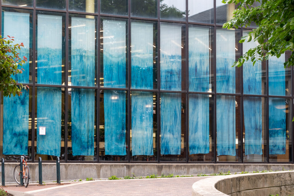 A translucent blue silk artwork hangs in an indoor gallery space, with layered cyan tones and organic patterns inspired by water and natural processes. The installation is displayed at Fletcher Free Library as part of Blue Alchemy by Renee Greenlee.